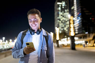 Lächelnder junger Mann mit drahtlosen Kopfhörern, der sein Smartphone in einer nächtlichen Stadt benutzt - WPEF06358