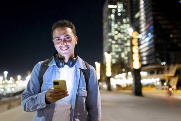 Happy man wearing wireless headphones holding mobile phone at night - WPEF06359