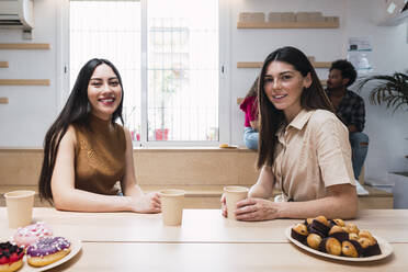 Lächelnde junge Geschäftsfrauen mit Einweg-Kaffeebechern sitzen am Tisch in einer Cafeteria - PNAF04466