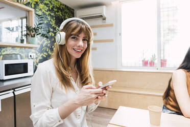Lächelnde Geschäftsfrau mit Kopfhörern, die mit ihrem Smartphone in einer Cafeteria sitzt - PNAF04467