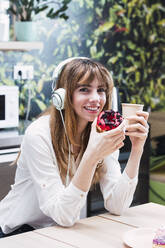 Smiling businesswoman wearing headphones sitting with disposable coffee cup and doughnut in cafeteria - PNAF04478