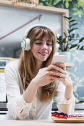 Smiling businesswoman with bangs text messaging through smart phone sitting in cafeteria - PNAF04480