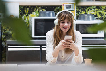 Lächelnde Geschäftsfrau mit Kopfhörern, die in einer Cafeteria über ihr Smartphone im Internet surft - PNAF04481