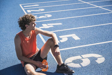 Sportsman holding water bottle sitting by starting line on track - UUF27145