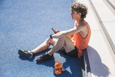 Young athlete using smart phone sitting by steps on sunny day - UUF27154