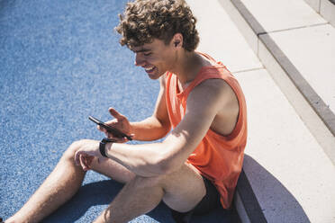 Glücklicher Sportler mit Smartphone vor einer Treppe sitzend - UUF27155