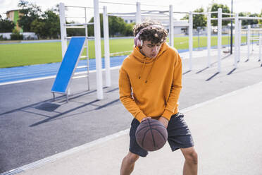 Young man listening music through wireless headphones and playing basketball - UUF27163