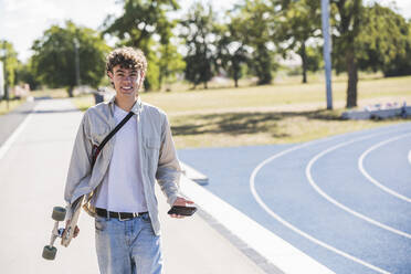 Happy young man holding skateboard and mobile phone on footpath - UUF27171