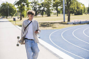 Lächelnder Mann mit Skateboard und Smartphone auf dem Gehweg - UUF27172