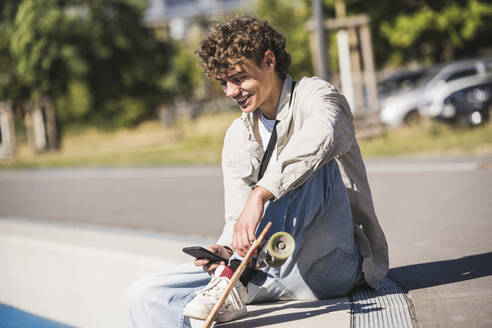 Glücklicher Mann mit Skateboard und Smartphone am Rande der Bahn sitzend - UUF27174