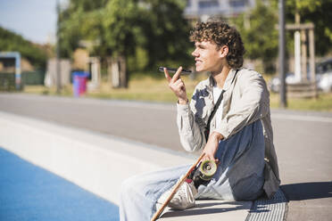 Mann mit Skateboard sendet Sprachnachricht über Smartphone am Rande einer Leichtathletikanlage - UUF27176