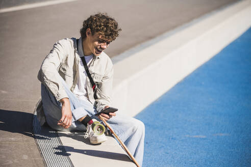 Fröhlicher Mann mit Skateboard und Smartphone am Rande einer Leichtathletikanlage - UUF27178