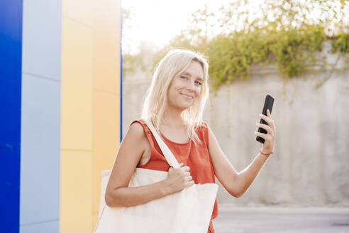 Lächelnde blonde Frau mit Mobiltelefon in der Hand an einem sonnigen Tag - EBBF05947