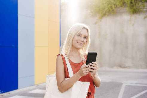 Lächelnde junge Frau mit blondem Haar hält Smartphone - EBBF05948