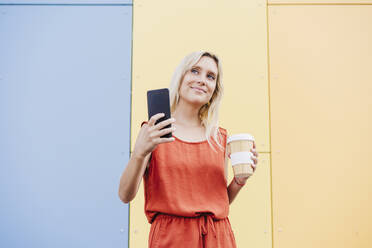 Lächelnde junge Frau mit Mobiltelefon und Einweg-Kaffeebecher vor einer bunten Wand - EBBF05950