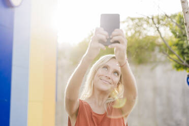 Lächelnde junge Frau macht ein Selfie mit ihrem Smartphone an einem sonnigen Tag - EBBF05951