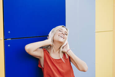 Happy woman enjoying music through wireless headphones in front of colorful wall - EBBF05971