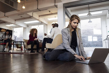 Young businesswoman using laptop sitting on office floor - WESTF25019