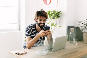 Happy freelancer using mobile phone sitting by laptop at home office - XLGF03057