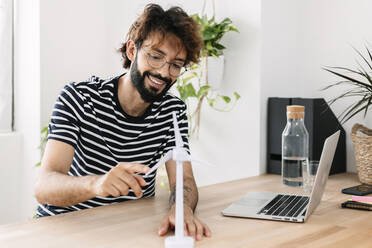 Happy freelancer touching wind turbine model by laptop at home office - XLGF03067