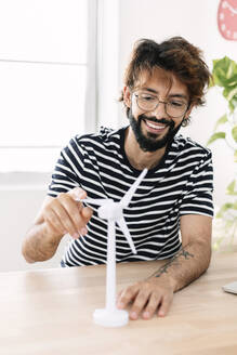 Happy freelancer sitting with wind turbine model at home office - XLGF03068