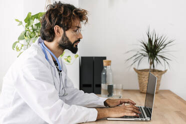 Doctor working on laptop sitting at desk in home office - XLGF03074