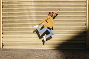 Happy young woman with hand raised jumping in front of shutter - ASGF02762