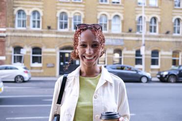 Happy young woman with curly hair holding disposable coffee cup - ASGF02766