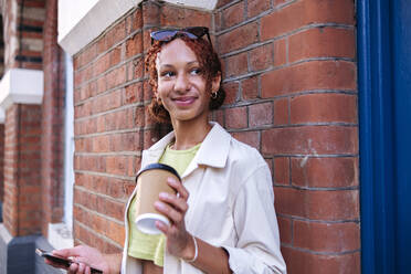 Lächelnde Frau hält Einweg-Kaffeebecher und Mobiltelefon an der Wand - ASGF02768