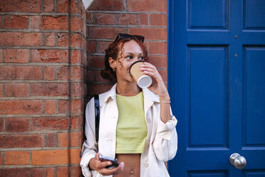 Junge Frau trinkt Kaffee aus einem Einwegbecher vor einer Wand - ASGF02769
