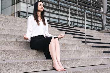 Contemplative woman sitting on staircase doing meditation outside office building - AMWF00389