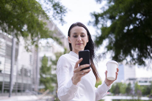 Mature businesswoman using smart phone and holding reusable cup - AMWF00391