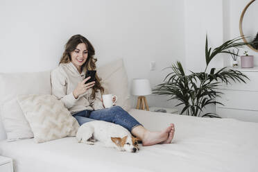 Junge Frau mit Kaffeetasse und Telefon, die neben einem Hund auf dem Bett zu Hause sitzt - EBBF05976