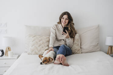 Lächelnde Frau, die über ihr Telefon im Internet surft und ihren Hund zu Hause streichelt - EBBF05978