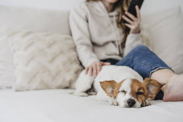 Frau streichelt Hund mit Telefon auf dem Bett sitzend zu Hause - EBBF05979