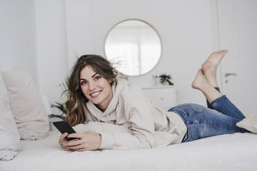 Lächelnde schöne junge Frau mit Mobiltelefon auf dem Bett liegend zu Hause - EBBF05993