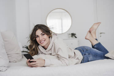 Smiling beautiful young woman with mobile phone lying on bed at home - EBBF05993