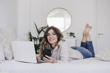 Nachdenkliche schöne Frau mit Laptop und Smartphone auf dem Bett liegend - EBBF05997