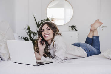 Smiling woman with laptop talking through phone lying on bed - EBBF06000