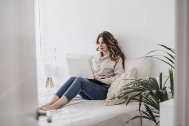 Junge Frau mit Laptop auf dem Bett sitzend zu Hause - EBBF06005