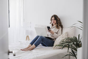 Junge Frau mit Laptop und Telefon auf dem Bett sitzend - EBBF06007