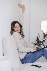 Young woman with laptop stretching on bed at home - EBBF06014