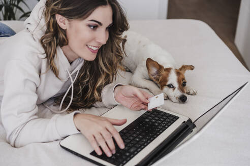 Young woman with credit card doing online shopping through laptop lying by dog at home - EBBF06021