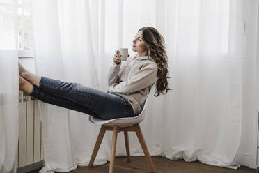 Young woman with coffee mug looking through window sitting on chair - EBBF06035