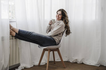 Happy woman with coffee mug sitting on chair at home - EBBF06036