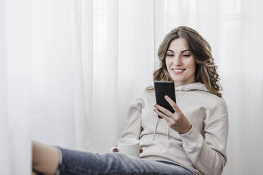 Smiling woman with cup using mobile phone at home - EBBF06038