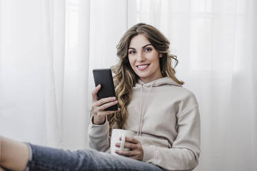 Smiling beautiful woman with coffee mug and mobile phone sitting at home - EBBF06039