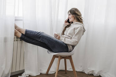 Smiling young woman talking over phone sitting on chair at home - EBBF06042