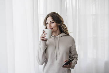 Frau mit Smartphone trinkt Kaffee bei weißem Vorhang zu Hause - EBBF06043