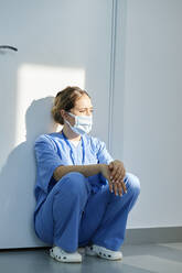 Krankenschwester mit Gesichtsschutzmaske hockt vor einer Wand im Krankenhaus - DSHF00531